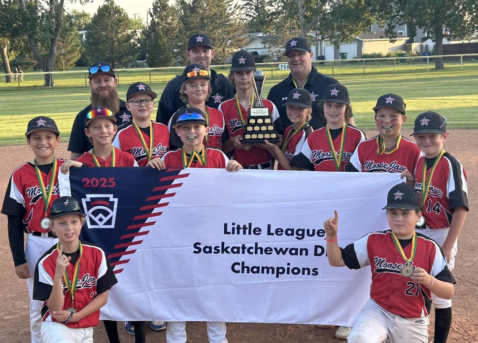A youth baseball team in red and black 'Moose Jaw' uniforms poses on a baseball field with their coaches. They hold a banner reading '2025 Little League Saskatchewan D1 Champions' and display a trophy. The players wear medals around their necks