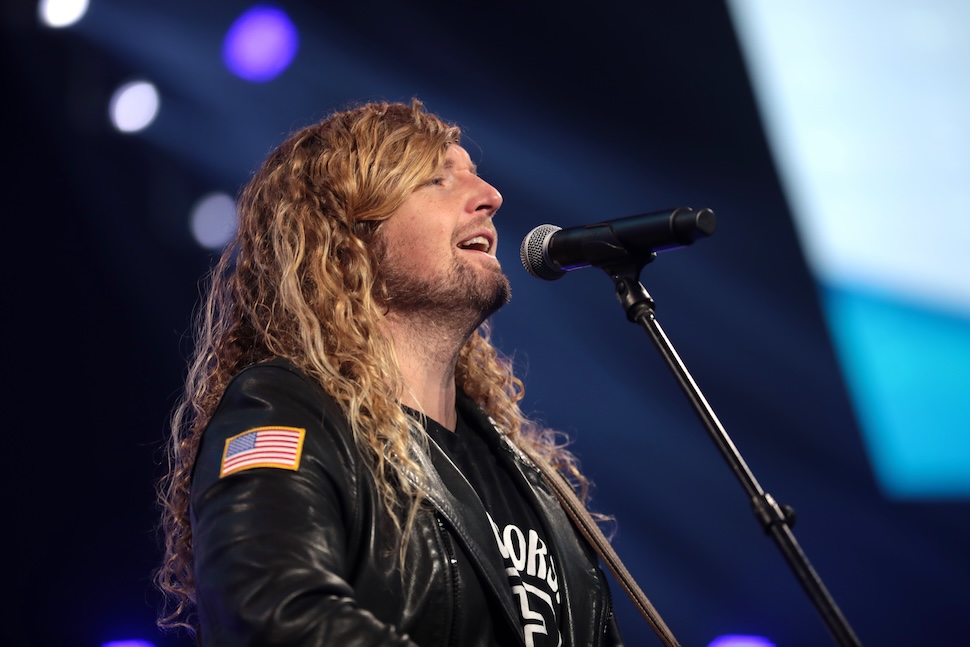 A man with long hair sings into a microphone on stage. he wears a black leather jacket with a USA flag shoulder patch