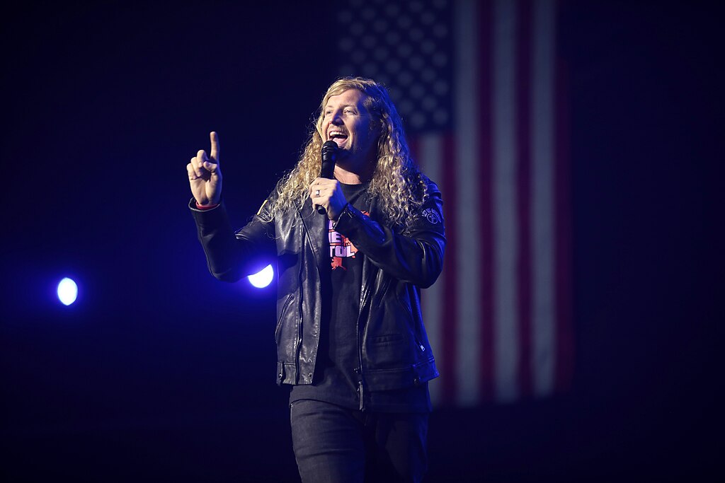 A man speaks from a stage, gesturing with one hand while holding a wireless microphone in another. A USA flag hangs behind him