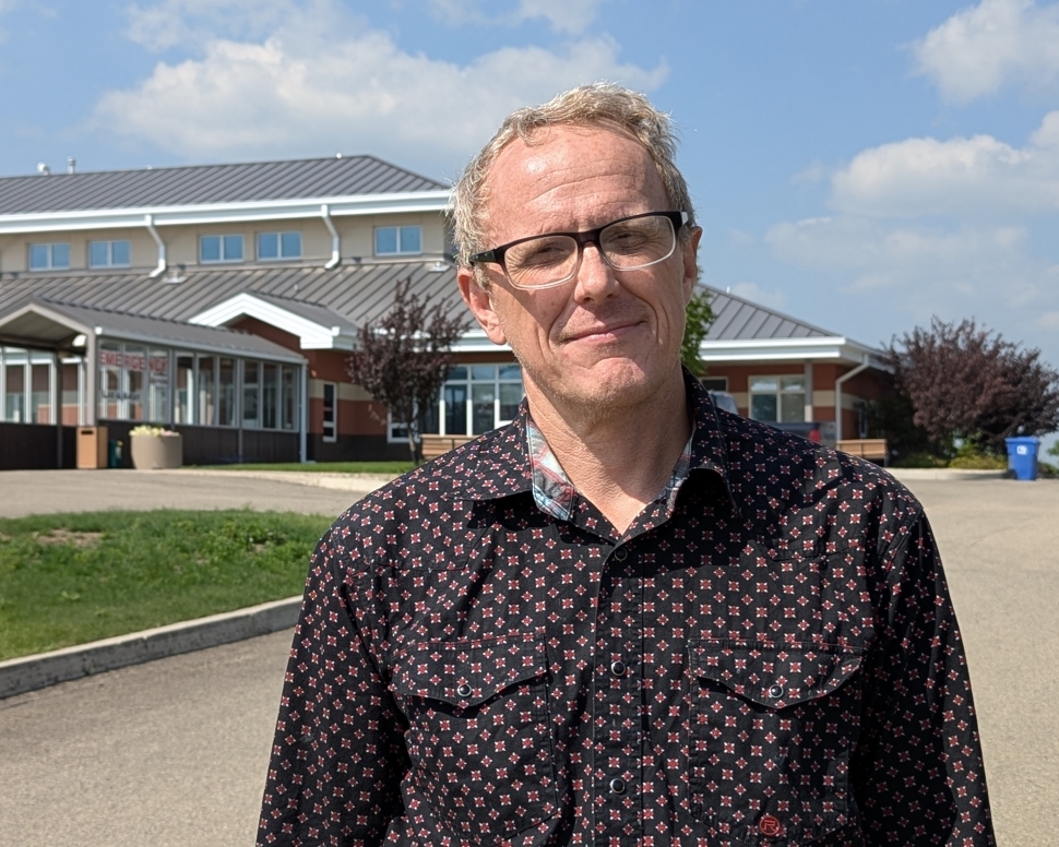 A man in a dark blue and red checked shirt standing in front of a brick building on a sunny day