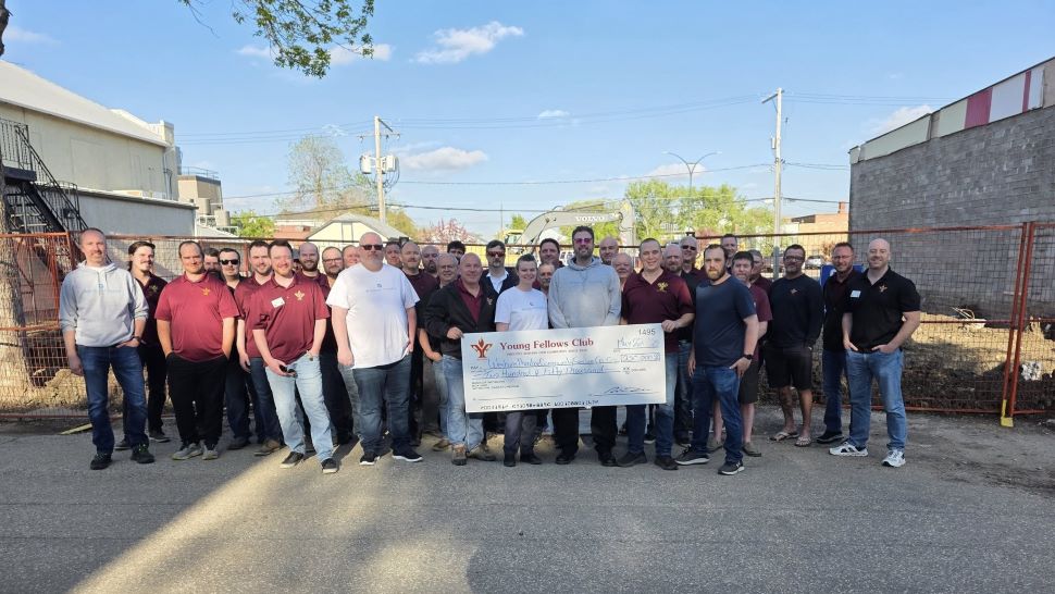 A group of men standing in front of a fenced off vacant lot, holding an oversized novelty cheque