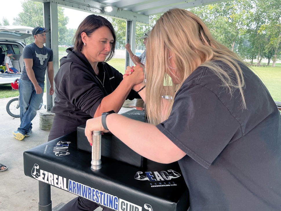 Two women locked in an arm wrestling match, smiling and focused, with a crowd cheering in the background.