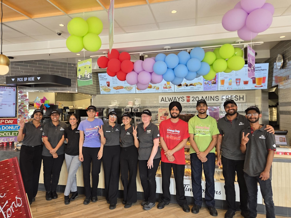 A group of smiling Tim Hortons staff members pose together inside the West End location in Portage la Prairie, wearing Camp Day shirts.