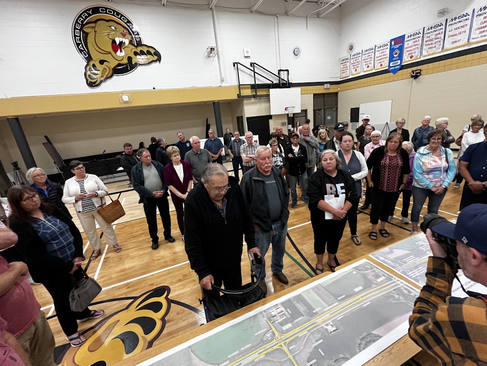 A large crowd of community members stands closely around a printed map of the proposed RCUT intersection design inside the Carberry Collegiate gymnasium on June 25, 2025.