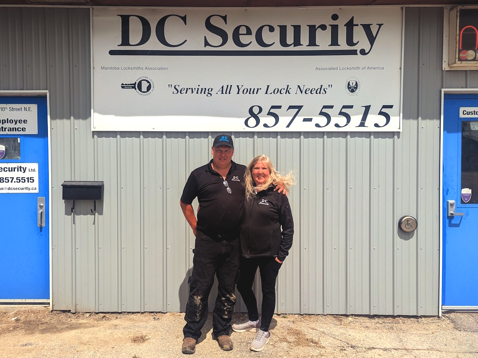 Dean and Angie Cairns stand smiling in front of the DC Security building, beneath a large overhead sign displaying the company name.