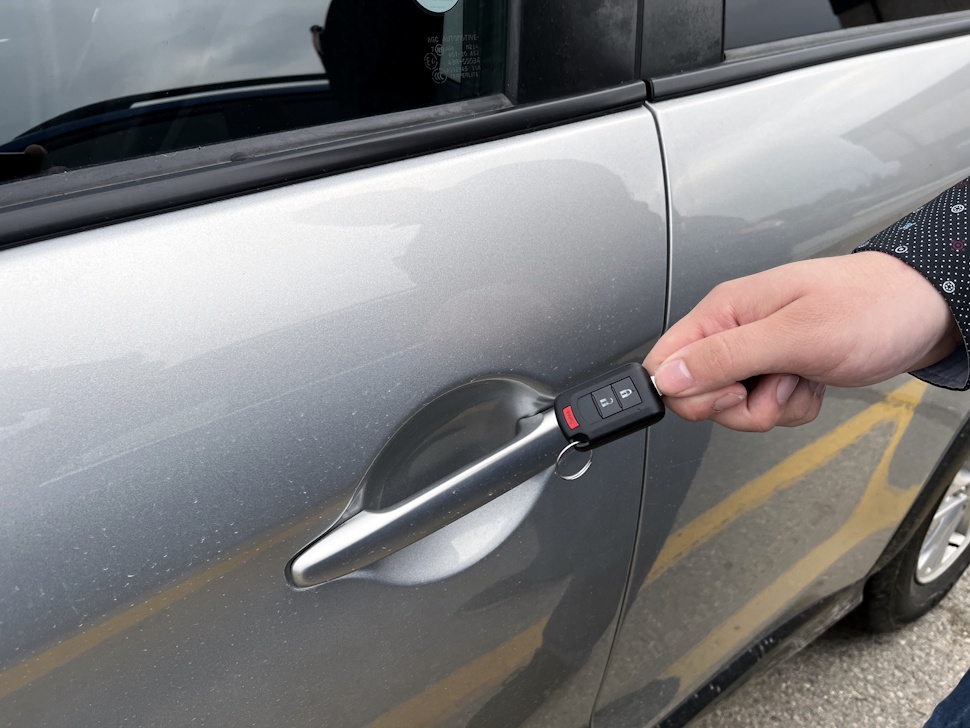 A person holds a vehicle key fob near the driver's side door of a parked car.