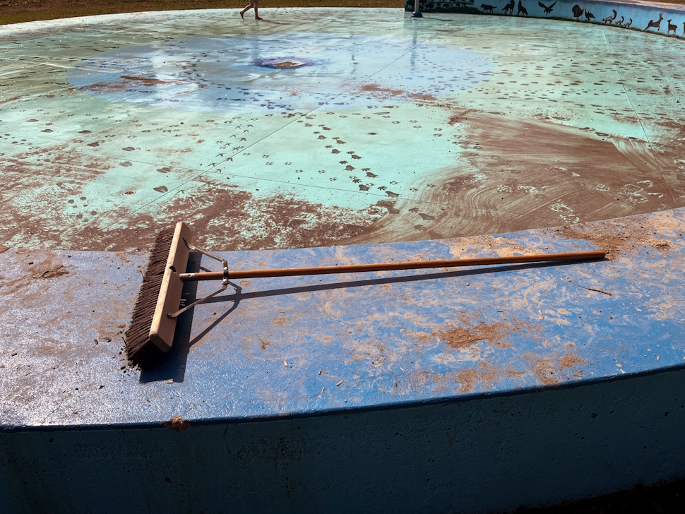 Mud and debris cover the surface of the Simplot Park splash pad, with a broom left nearby during cleanup efforts.
