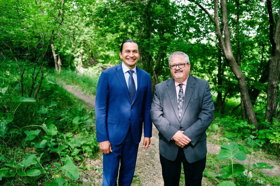 two men standing in a wooded forest smiling at the camera