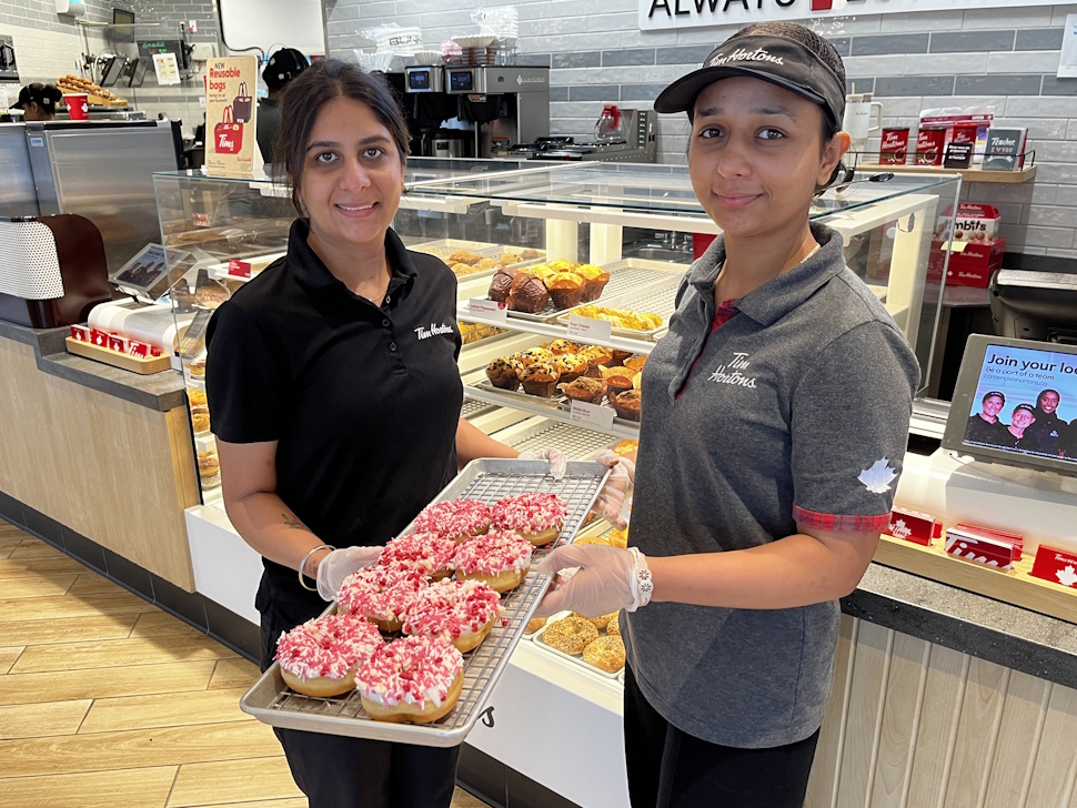 Simren Kaur and Manpreet Kaur smiling and holding a tray of Camp Day donuts inside the West End Tim Hortons.