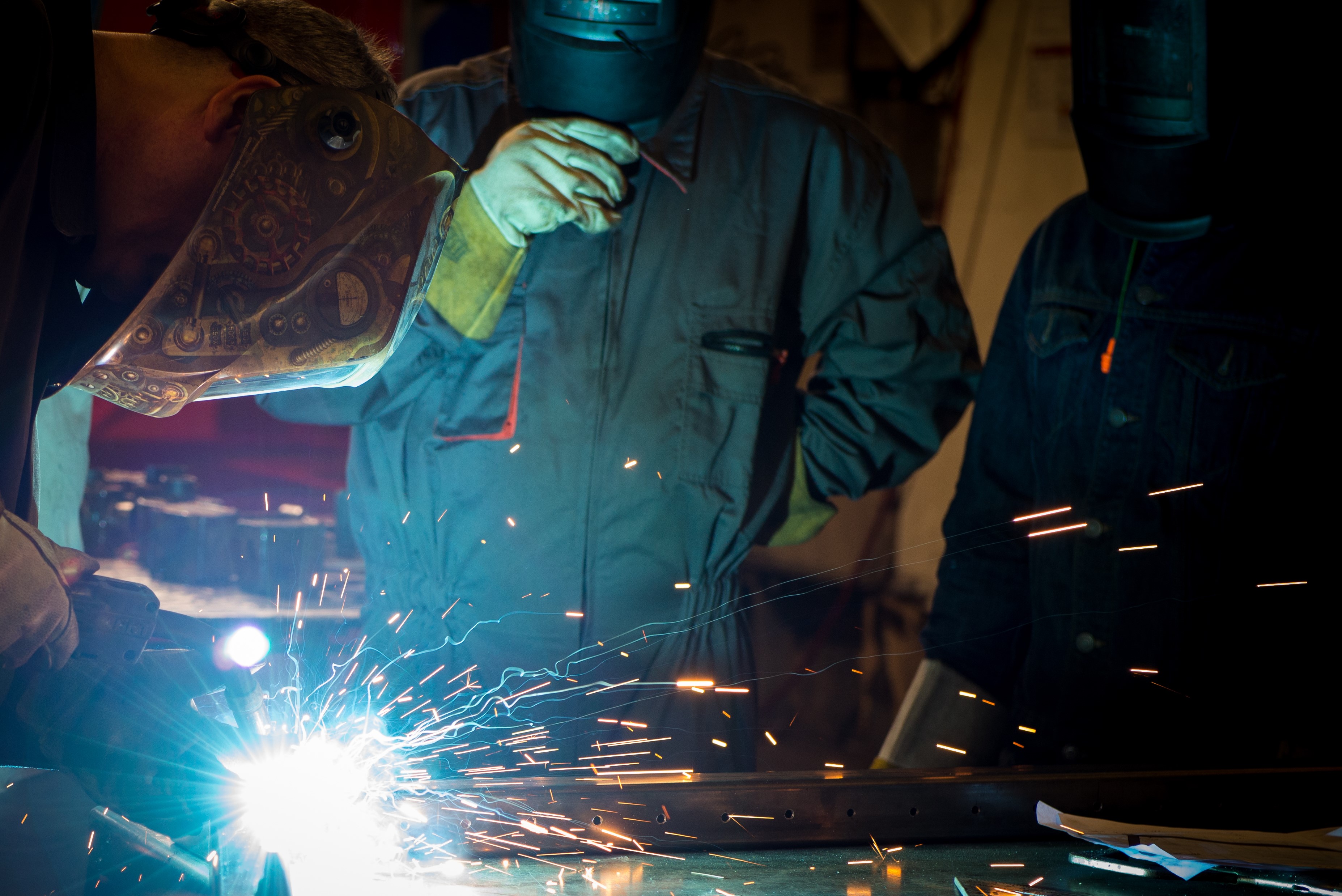 Sparks fly as a person welds a piece of metal. 2 other people are seen watching while using welding helmets