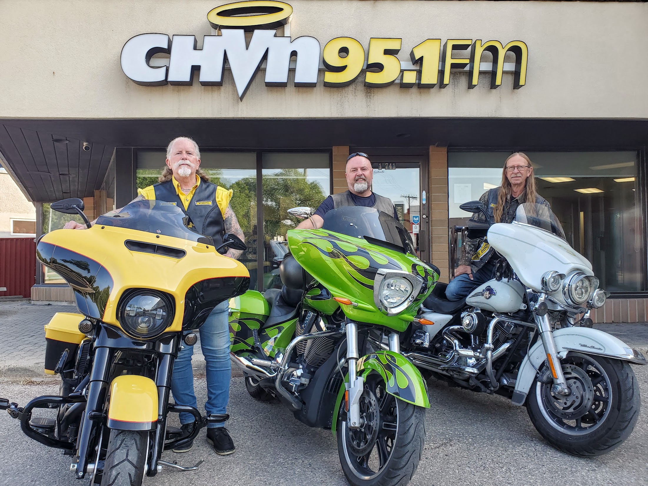 3 men stand beside 3 motorcycles parked under a CHVN logo sign outside a building