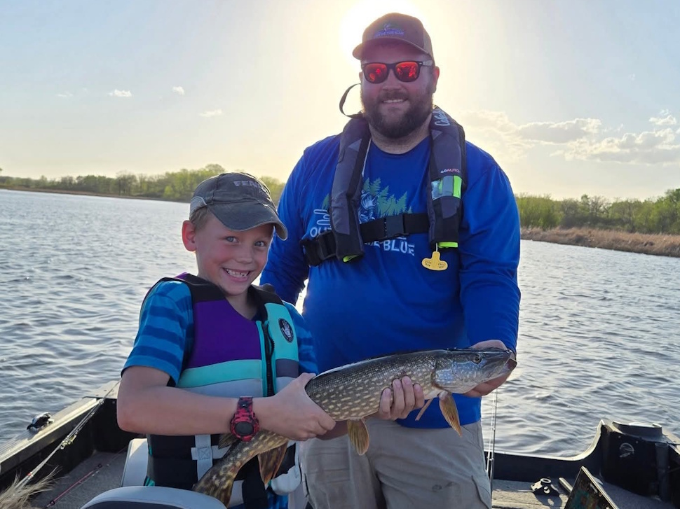 A man and a boy proudly holding up a fish they caught while fishing by a calm lake.