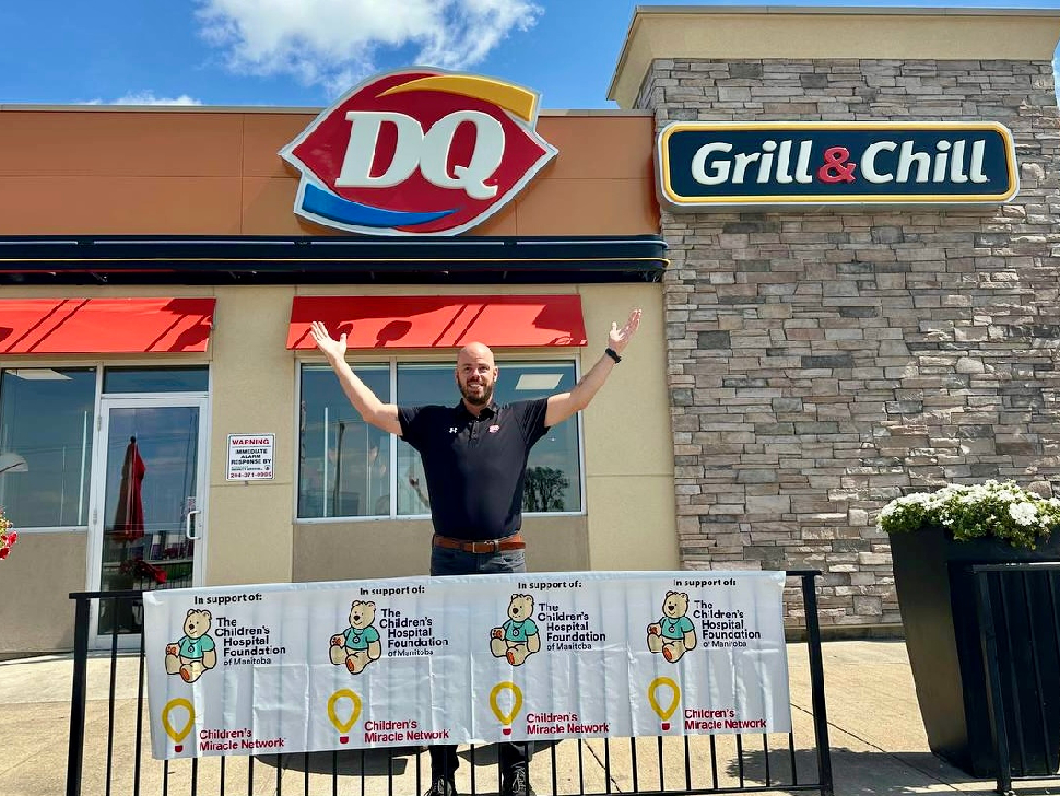 Stewart Holditch stands with his hands in the air, looking excited, in front of Dairy Queen in Steinbach. A promotional banner for Miracle Treat Day and the Children's Hospital Foundation is in front of him.