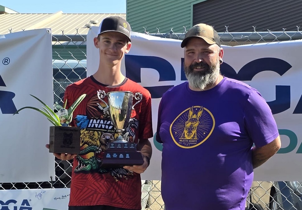Two individuals standing in front of a PDGA banner. The person on the left wears a red 'INNOVA DISC GOLF' shirt and holds a trophy and a potted plant. The person on the right wears a purple 'MAJESTIC ASAMATS' shirt featuring an illustrated animal head with sunglasses