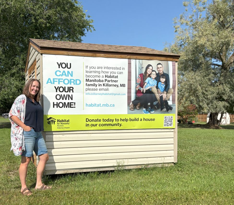 a woman standing beside a shed with a poster on it for Habitat for Humanity Manitoba