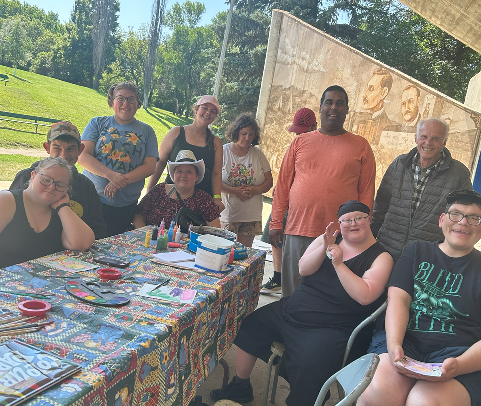 A group of people gathered around a table outdoors, covered with a colorful tablecloth and art supplies like paint palettes, brushes, and bottles. Trees and greenery surround the area, with a mural of historical figures visible in the background.
