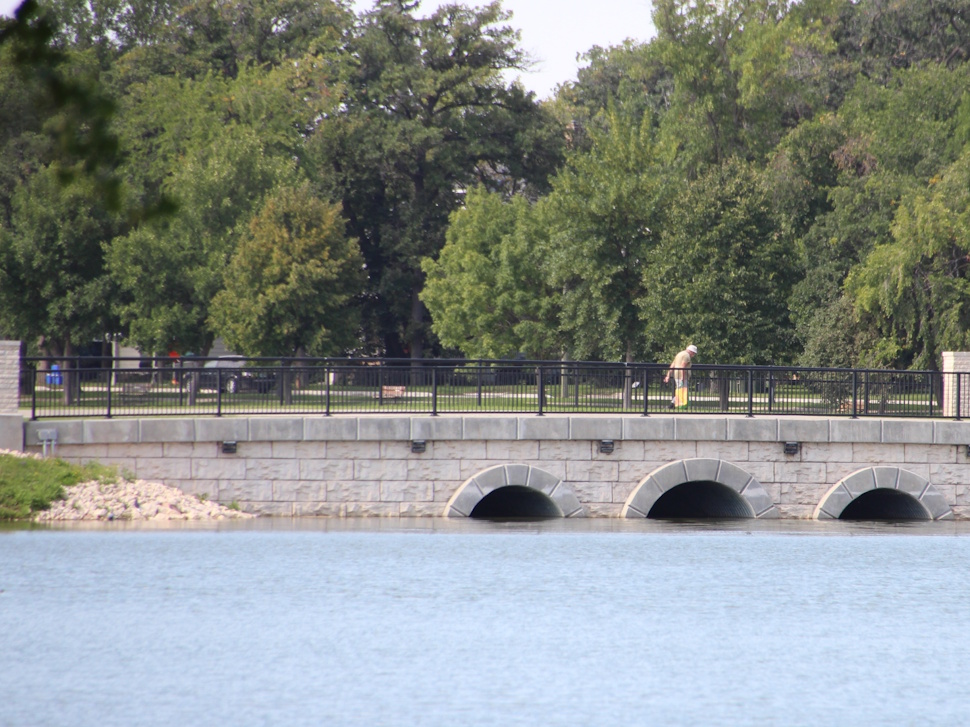 Crescent Lake in Portage la Prairie on a sunny summer day, with calm water, grassy shoreline, and trees in the background.