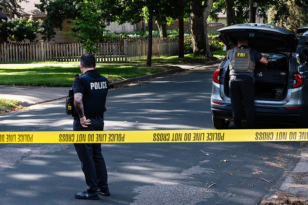 A police officer stands on a street behind yellow police tape. An FBI agent is seen removing items from the back of an SUV
