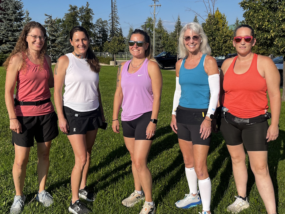 Five women from the Positive Portage Runners stand together smiling in front of Stride Place in Portage la Prairie. From left to right: Diane Wing, Vanessa Taylor, Stacey Moffat, Amber Driedger, and Vanessa Schurko.