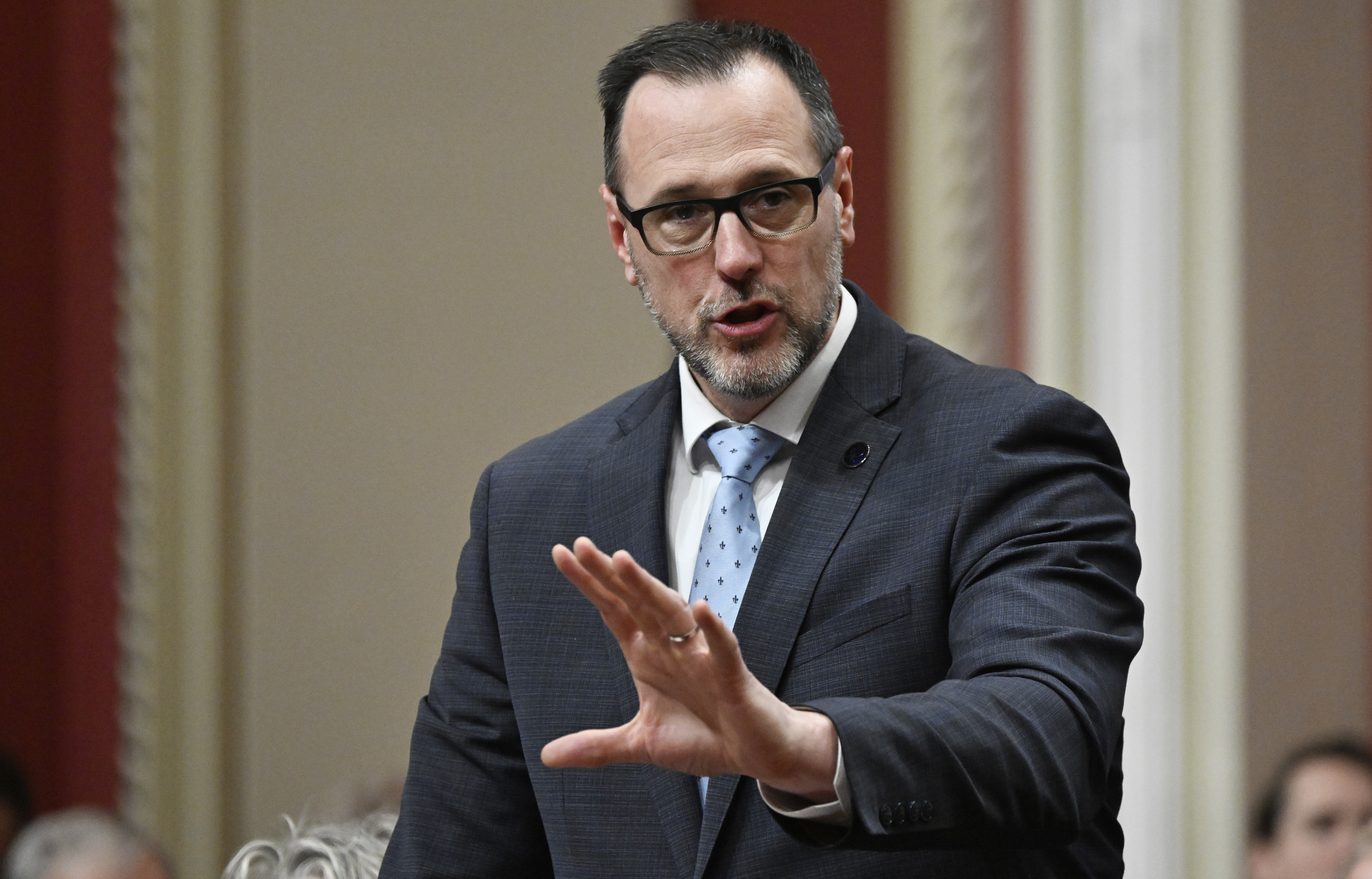 A man stands and speaks while gesturing with his left hand
