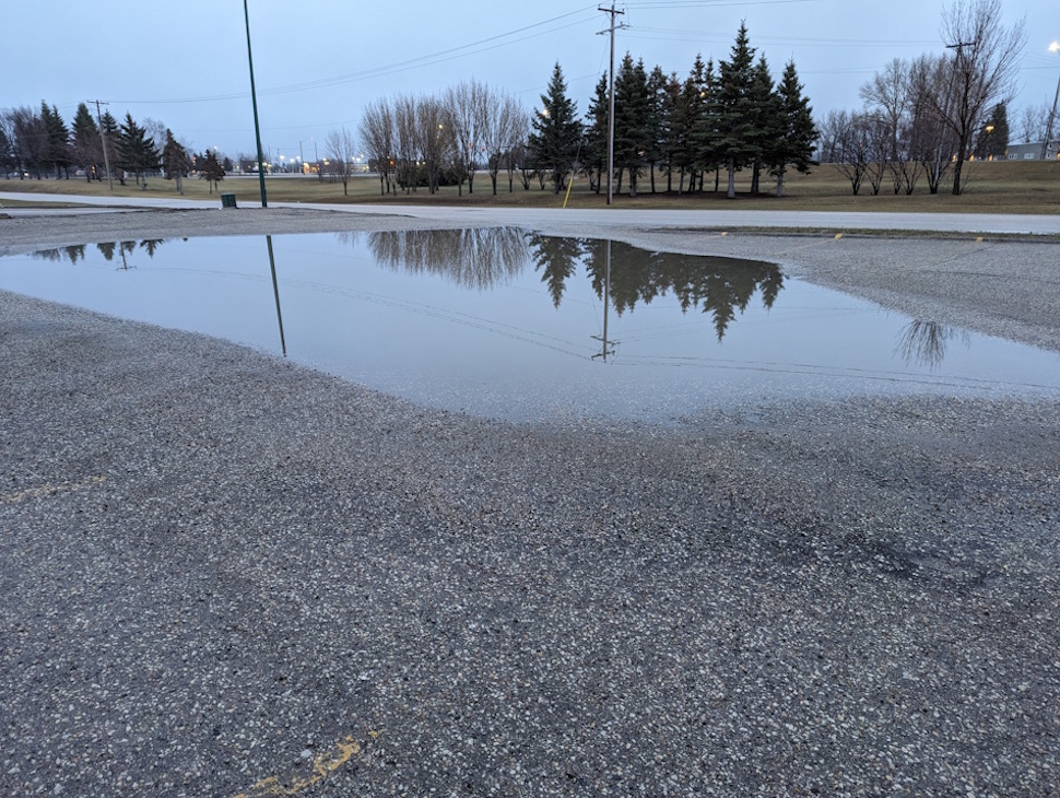 A large rain puddle reflects grey skies in Portage la Prairie on Friday morning, as wet weather and smoky air continue to hang over much of southern Manitoba. PortageOnline/File Photo