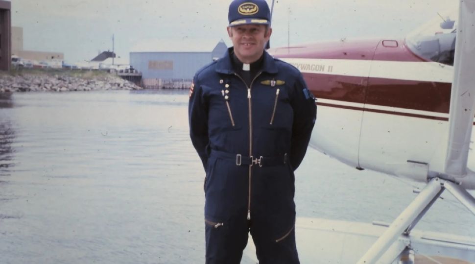 A man dressed in a blue flight suit stands in front of a float plane. His clergy collar can be seen below the suit