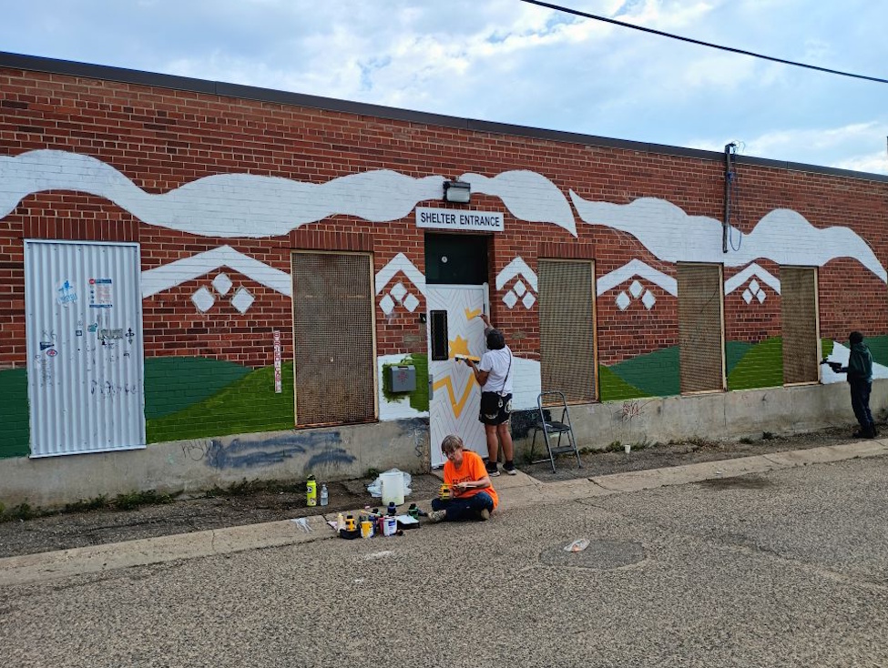 two people painting an outdoor mural on a red brick wall