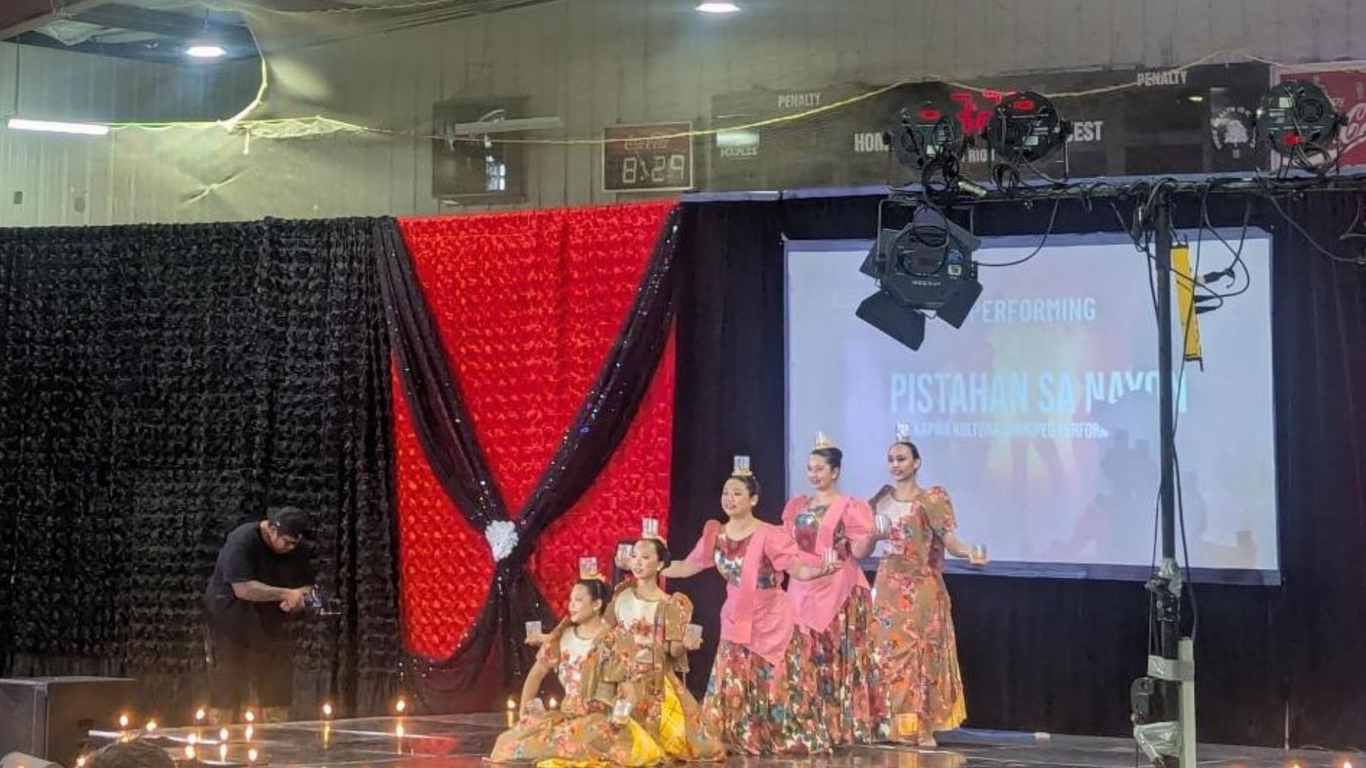 Dance performers on stage at a Filipino festival. The dancers are in colourful flowery attire and are holding cups in their hands and have them on their heads.