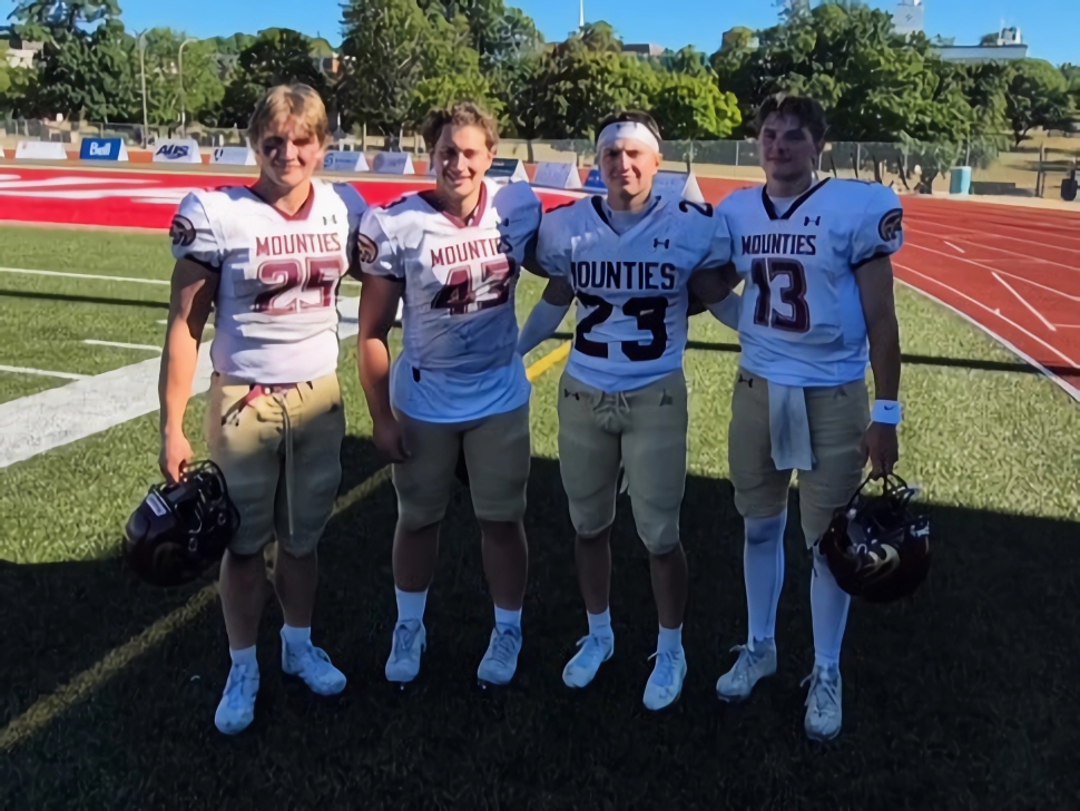 Four football players in white jerseys and light gold pants with red letters and numbers