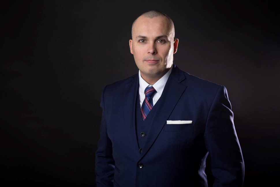 A man in a dark suit standing in front of a dark background. 