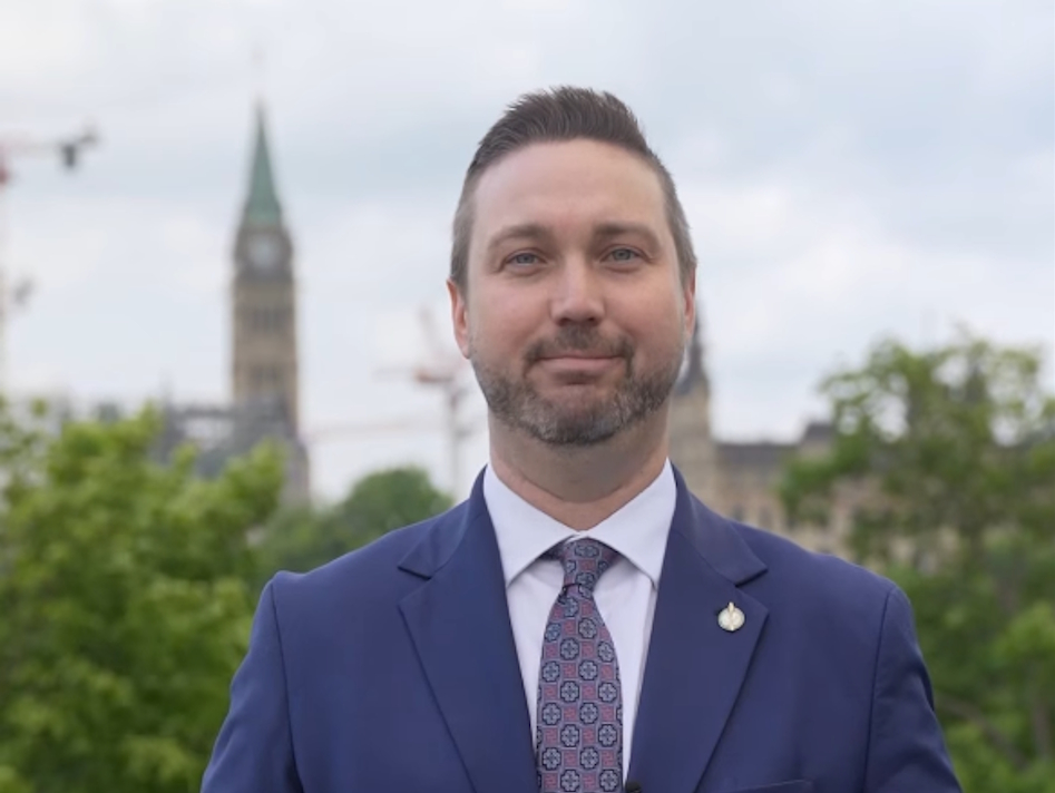 Branden Leslie in Ottawa standing before the Parliament building peaking above trees immediately behind him