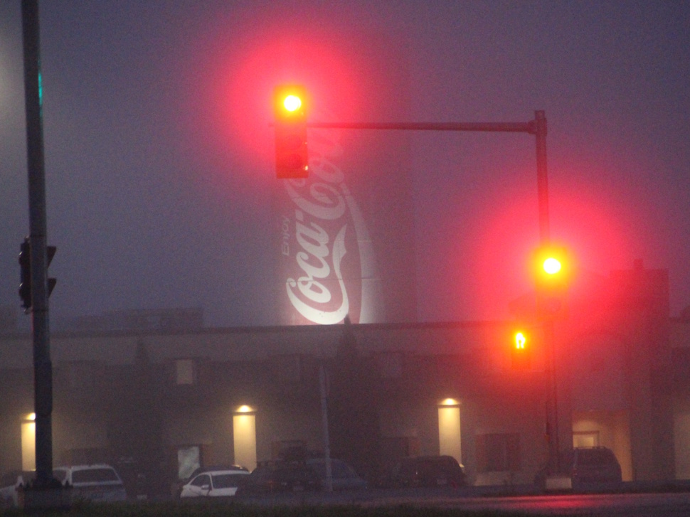 A foggy morning in Portage la Prairie with traffic lights glowing red and yellow through the mist, and a large Coca-Cola sign faintly visible in the background.