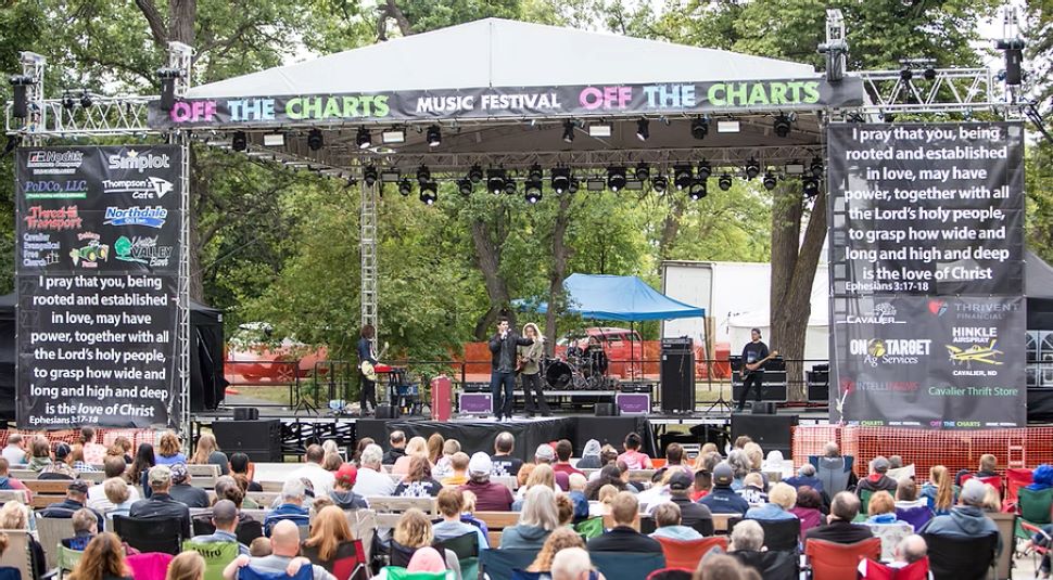 A crowd sits in lawn chairs and watches a speaker on an outdoor stage