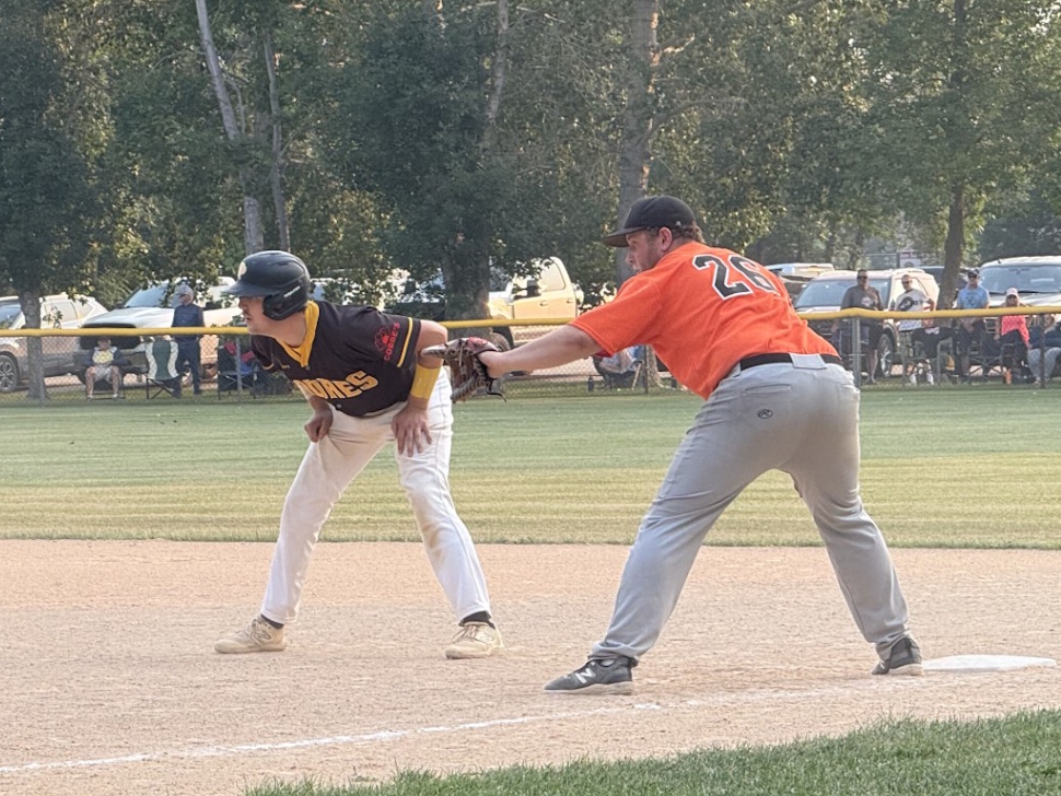 A Portage Padres player leads off first base as a Plumas Pirates fielder prepares to catch a throw from the pitcher for a pickoff attempt.