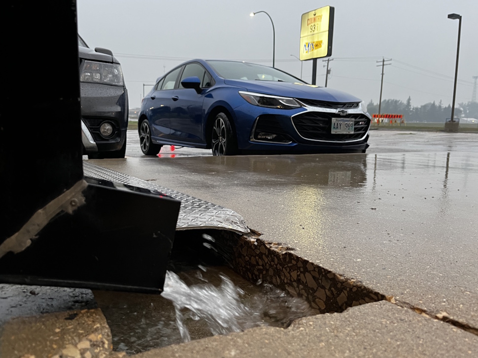 Water flows rapidly out of a parking lot drain onto a wet concrete surface during heavy rain, with a blue car and a cloudy, rainy sky in the background.