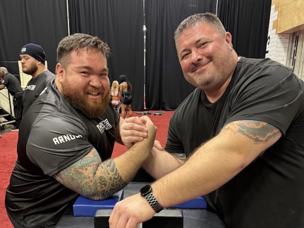 Ryan Espey smiles at the camera while arm wrestling an opponent at a competition table.