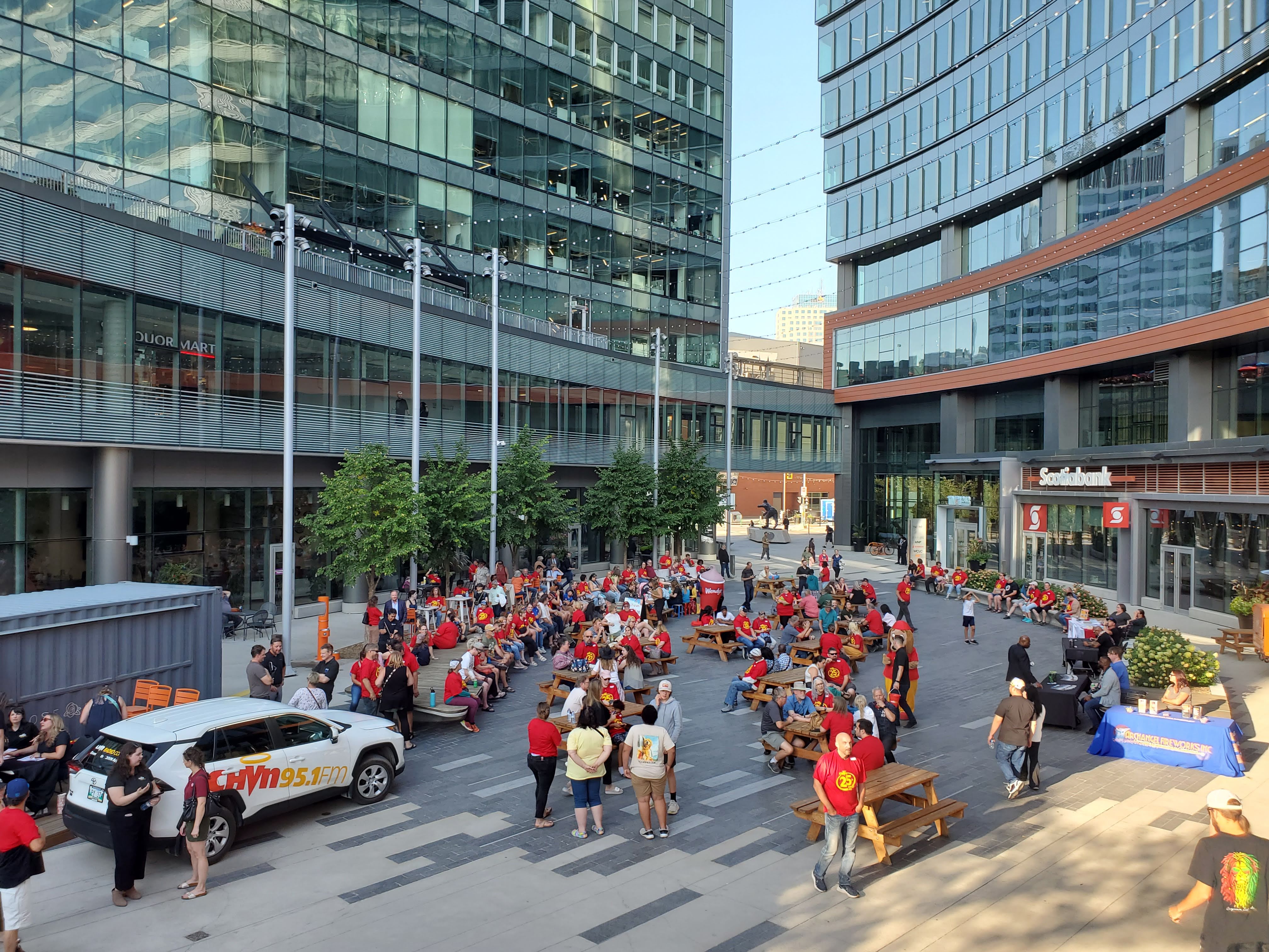 A HIGH AND WIDE ANGLE SHOT OF A LARGE CROWD SPREAD THROUGHOUT TRUE NORTH SQUARE VISITING AND SITTING AT PICNIC TABLES