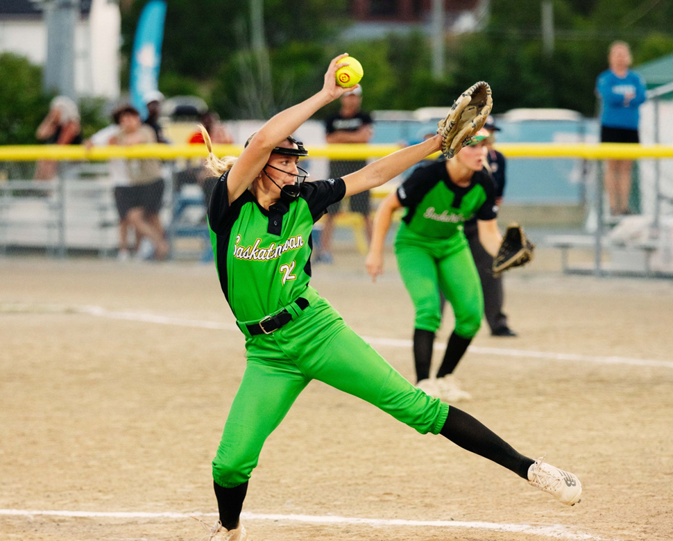 Softball player in a green and black "Saskatchewan" uniform mid-pitch, holding a yellow ball with one arm raised and the other extended forward with a glove; another player and spectators are visible in the background behind a yellow barrier.