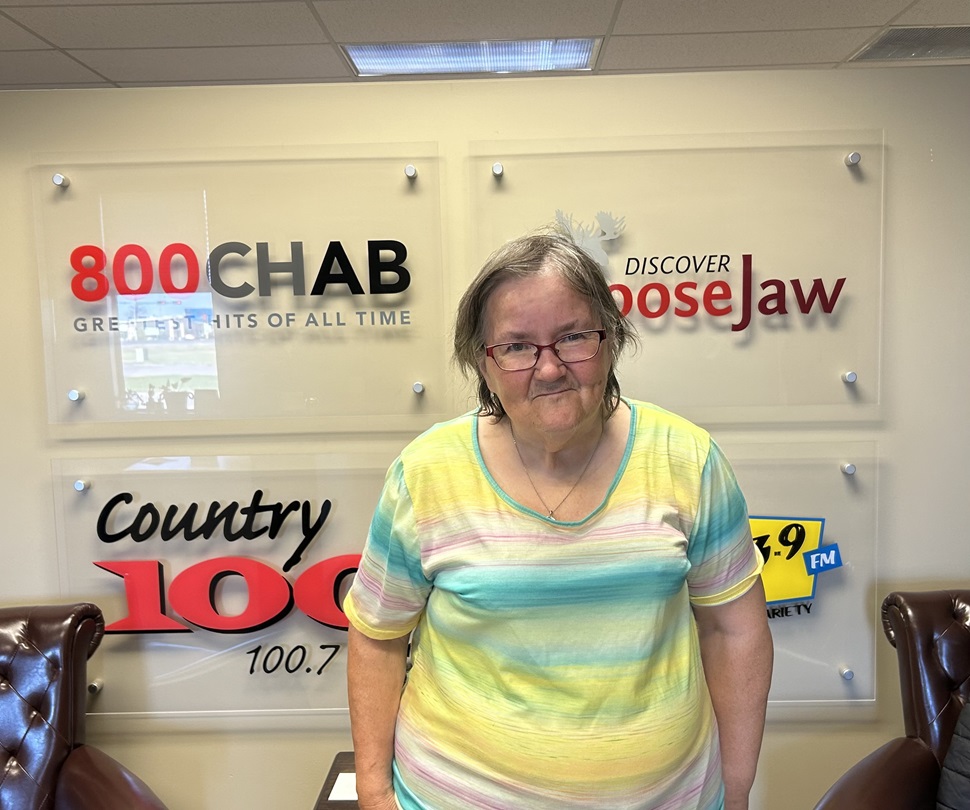 A woman wearing a striped pastel shirt stands indoors in front of a wall with radio station logos, including 800 CHAB, Country 100, and Mix 103.9 FM, along with the Discover Moose Jaw logo.