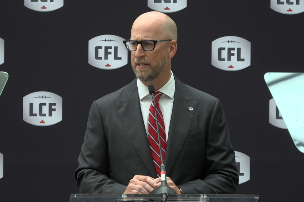 A man stands behind a podium and speaks into a microphone. a black backdrop has the cfl logo on it