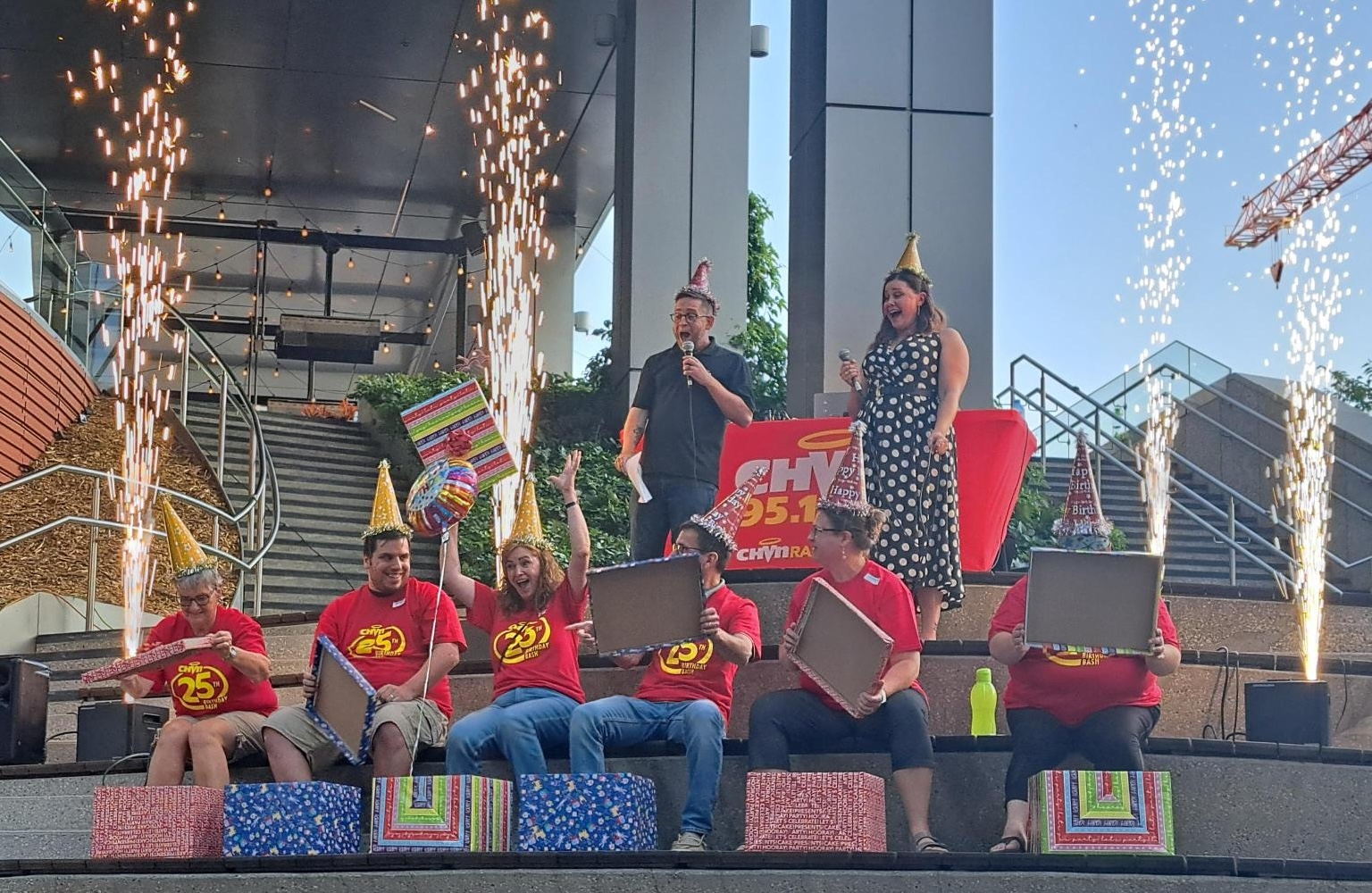 6 people wear matching "chvn 25th birthday bash" red tshirts and sit on steps beside each other. Large wrapped boxes sit in front of each person and 1 woman raises her arms as a balloon floats in front of her. 4 large sparklers shoot into the air and 2 events hosts smile