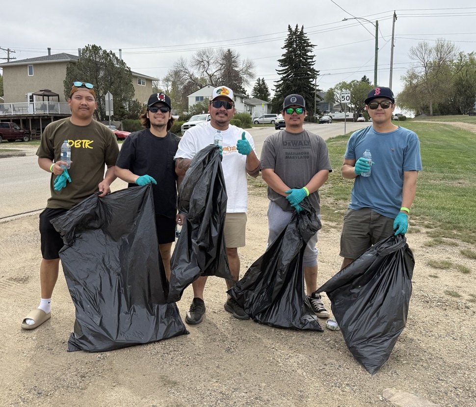 Five people stand on a dirt path holding large black garbage bags and wearing gloves, participating in a community cleanup. Residential houses, trees, and a 50 km/h speed limit sign are visible in the background.