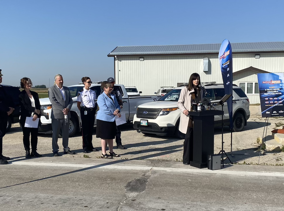 A woman speaks at a podium outside on the side of a road while a group of other dignitaries stand behind and listen