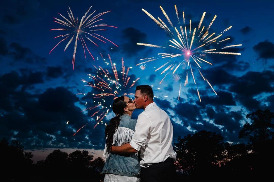 A man and woman kiss under the night sky as fireworks go off overhead