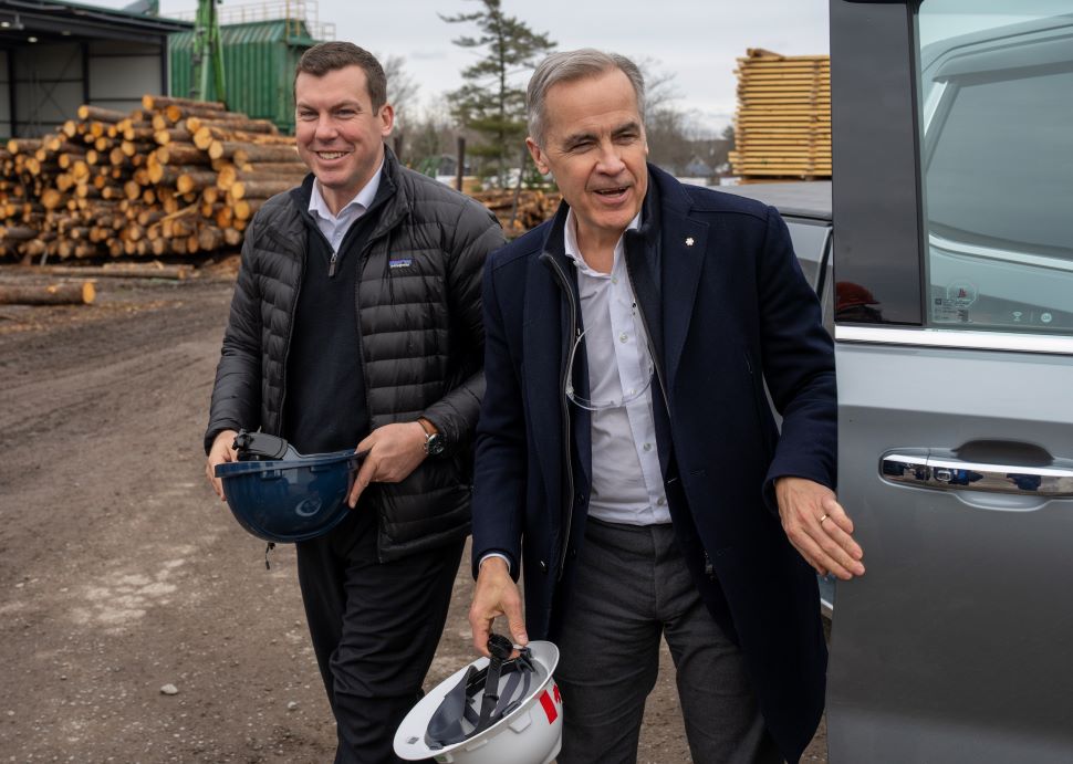 2 men smile holding hard hats as they exit a vehicle and stand in a parking lot. Stacks of logs are seen in the background.