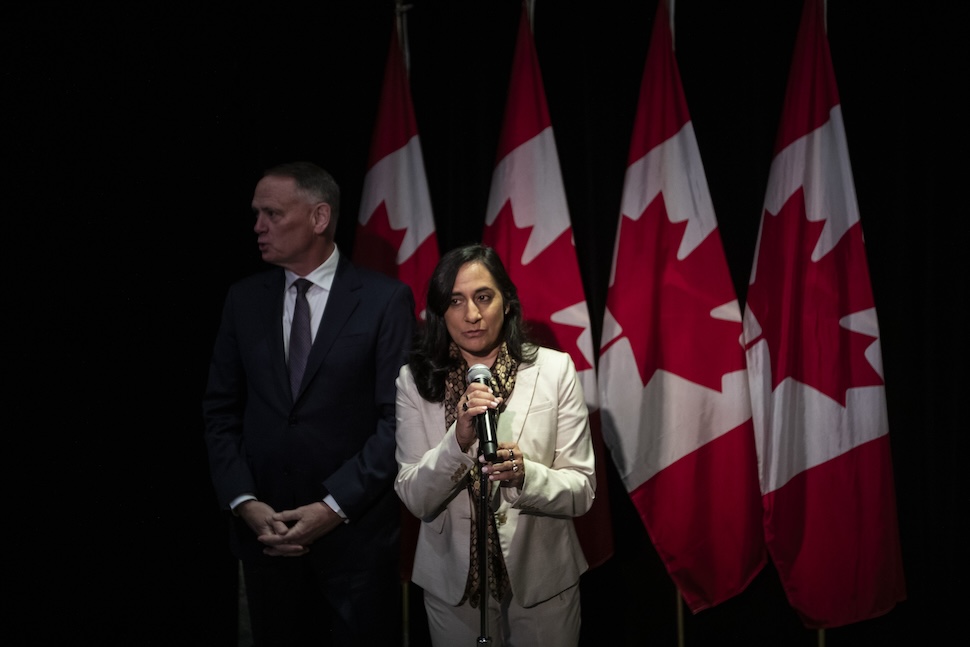 A woman speaks into a microphone while a man stands behind her and looks on. There are 4 Canadian flags draped from poles behind them