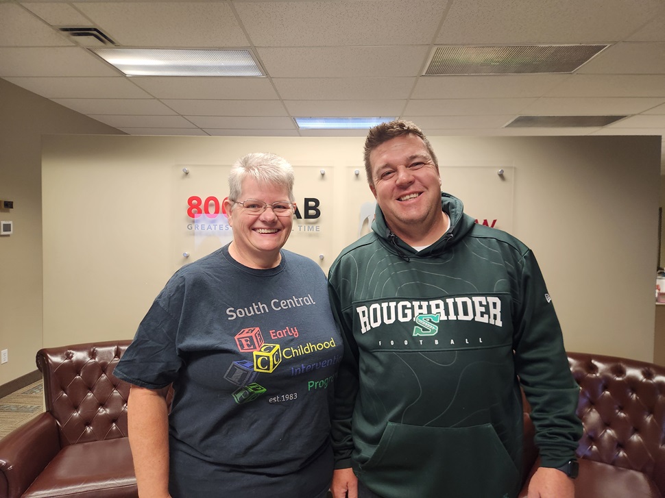 Two people smiling while standing side by side in a radio station lobby. One is wearing a South Central Early Childhood Intervention Program T-shirt, and the other is wearing a Saskatchewan Roughriders hoodie.