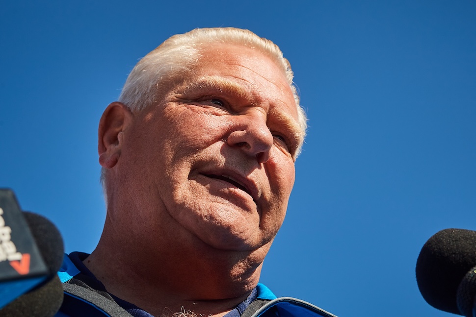 A low-angle of Doug Ford speaking to reporters with microphones in the bottom edges of the photo