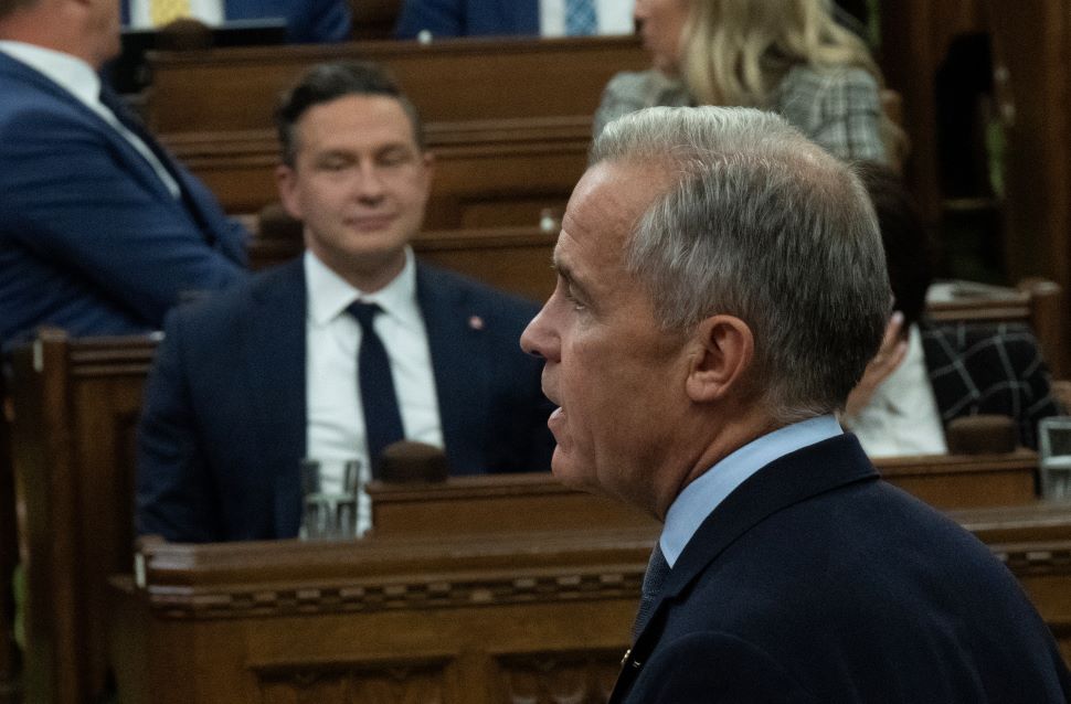 A picture from behind of Mark Carney standing and speaking in the House of Commons. Pierre Poilievre can be seen in the background seated across the aisle. 