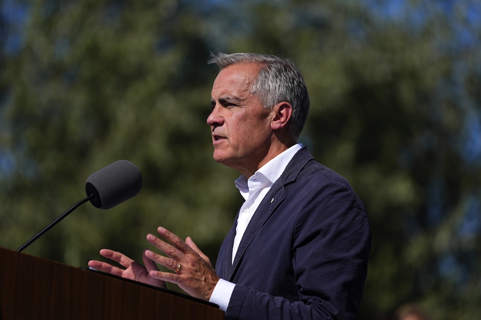A side angle shot of Mark Carney speaking behind a podium. He is outdoors with trees in the background, and gestures with his hands while talking